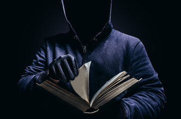 Photo of scary shaded hooded man holding book and turning the pages on dark background. Horror book concept.
