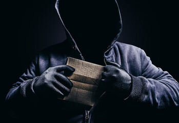 Photo of scary shaded hooded man hiding cardboard box parcel on dark background.