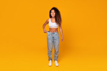 african lady showing slim waist in oversized jeans, yellow background