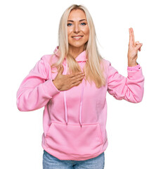 Young blonde woman wearing casual sweatshirt smiling swearing with hand on chest and fingers up,...