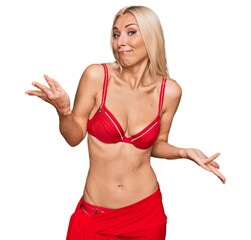 Young blonde woman wearing bikini clueless and confused expression with arms and hands raised....