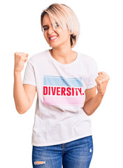 Young beautiful blonde woman wearing t shirt with diversity word message very happy and excited doing winner gesture with arms raised, smiling and screaming for success. celebration concept.