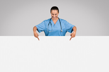 Lady nurse in blue medical uniform pointing down at white placard board against grey background, banner