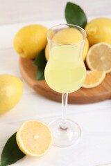 Liqueur glass with tasty limoncello, lemons and green leaves on white wooden table, closeup