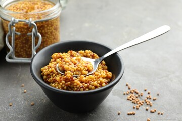 Whole grain mustard in bowl, spoon and dry seeds on grey table