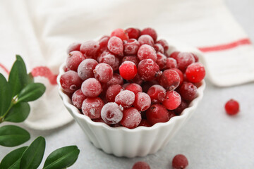 Frozen red cranberries in bowl and green leaves on light table, closeup