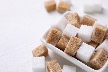 Different sugar cubes in bowl on white table, closeup. Space for text