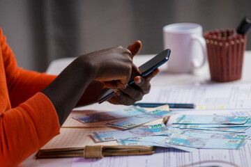 Black person in orange long sleeves shirt working on desk, writing in notebook and holding mobile phone with Brazilian real notes on desk. accounting concept
