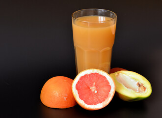 A mixture of fruit juices in a tall glass on a black background, next to pieces of ripe grapefruit and mango.