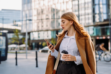 Young positive businesswoman using mobile phone while standing on sunny street near office building