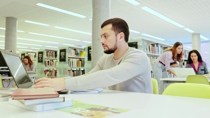 Focused guy student preparing for an exam on a laptop in the university library, looking for sources of information in textbooks. High quality 4k footage
