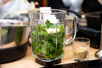 Fresh Greens and Garlic in Blender for Pesto. Preparation for pesto sauce with fresh basil leaves, garlic cloves in a glass blender jar on a kitchen countertop, pine nuts in the background.