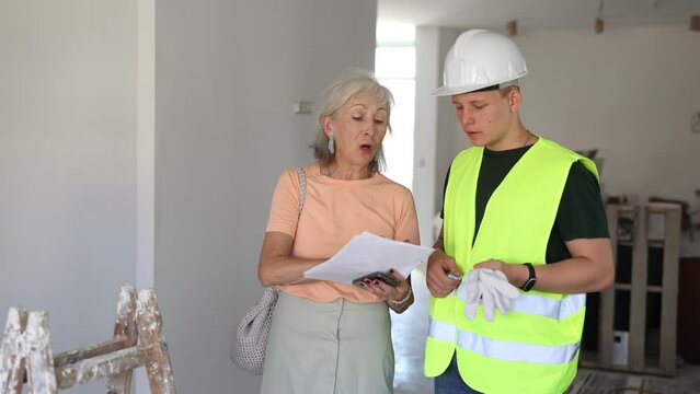 Portrait of mature woman architect talking with teenager construction worker in apartment under reconstruction, examining documents and planning work process for renovation