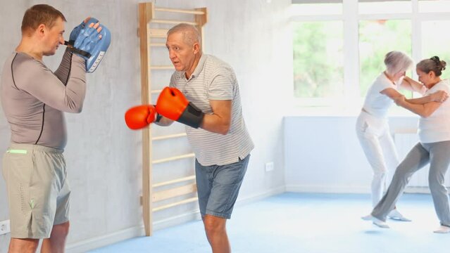 Diligent old man training boxing kicks on punch mitts held by instructor in sports hall. High quality 4k footage
