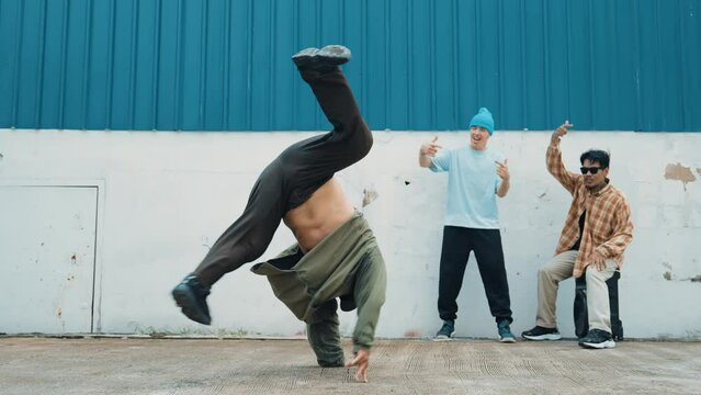 Skilled hipster doing cart wheel while multicultural friends encourage him. Hip-hop dancer moving foot step to music surrounded by group of hipster at wall near street. Outdoor sport 2024. hiphop.