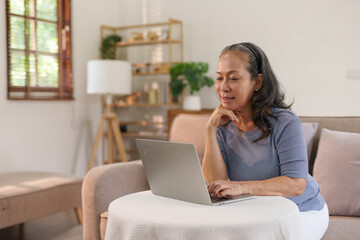 Senior asian woman sitting on sofa using laptop computer, working, chatting, social media internet at home.