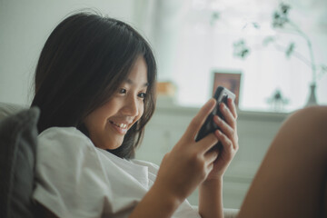 Happy Asian girl relaxing on a comfortable couch at home texting and messaging on a smartphone,...