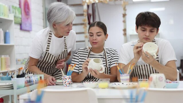 Positive enthusiastic senior woman offering guidance to interested young couple, girl and guy, painting ceramic mugs in pottery class. Creative hobby concept
