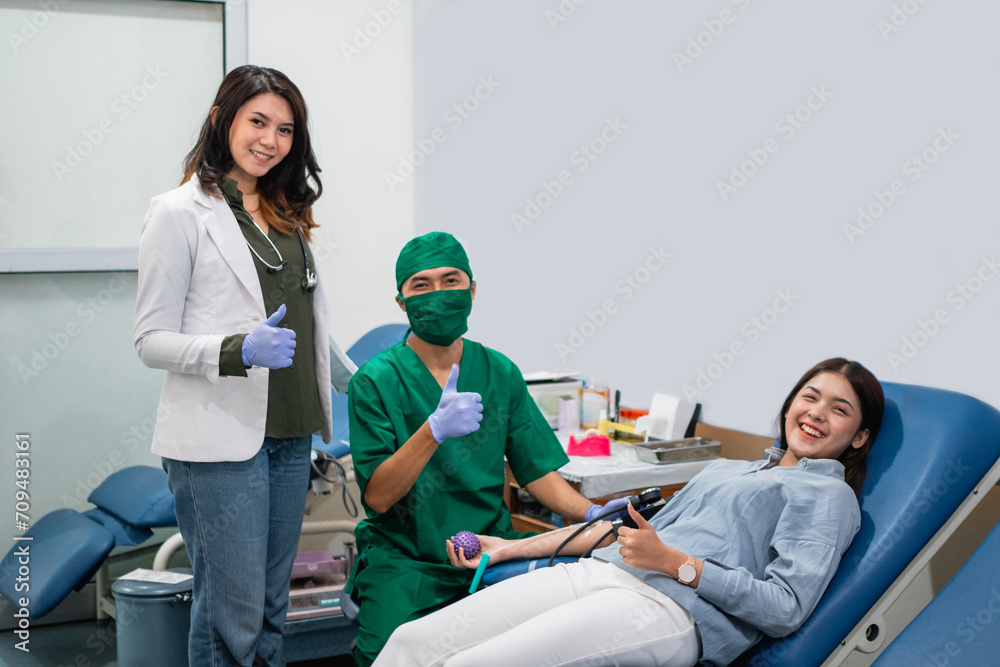 Wall mural woman donor and two doctors smile at the camera with thumbs up in the blood transfusion room