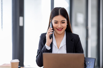 Asian Woman entrepreneur busy with her work in the office. Young Asian woman talking over smartphone or cellphone while working on computer at her desk.
