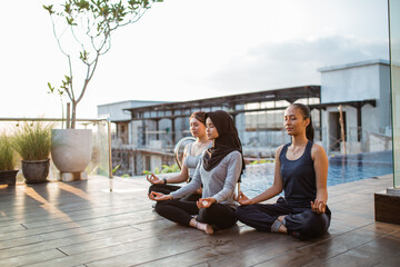 three young woman sitting near swimming pool in lotus position at resort, closed eyes, meditation
