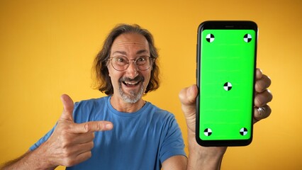 Excited mature elderly man standing and pointing Big smartphone with blank green screen with tracking points, demonstrating copy space for app or website design, mockup image on yellow background