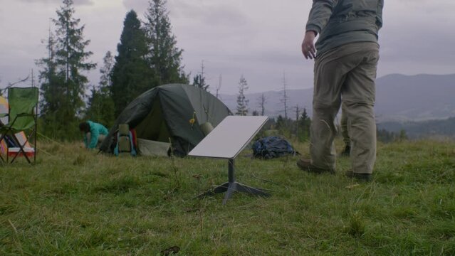 Group of hikers set up campsite in mountains. Caucasian man with son sets up and connects antenna of satellite internet in camp. Hikers rest during their vacation to mountains. Tourism and travel.