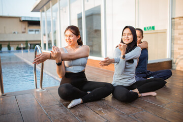 Obraz premium three young women sitting crosslegged doing arm stretches near a hotel swimming pool