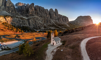 South Tyrol, Italy - Aerial panoramic view of the Chapel of San Maurizio (Cappella Di San Maurizio) at the Passo Gardena Pass in the Italian Dolomites at autumn with clear blue sky and setting sun - obrazy, fototapety, plakaty