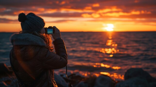  a woman taking a picture of the sun setting over the ocean with a camera in front of her, with the sun setting over the water behind her.
