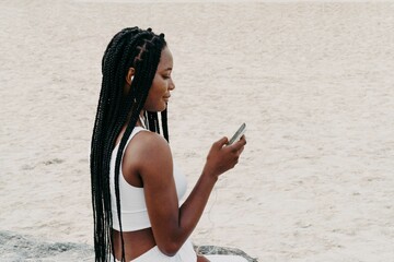 Woman with braids with headphones sitting at the beach using phone