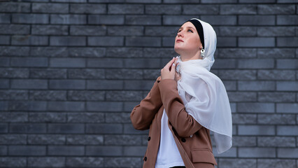 A young blue-eyed woman in a hijab looks up with her arms folded across her chest against a gray brick wall.