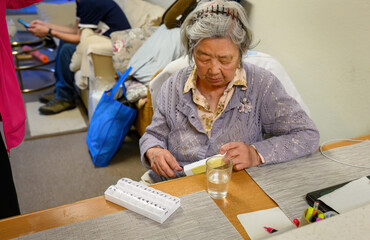 An elderly woman checking her medication in the pill box at home. Family members in the background. California.