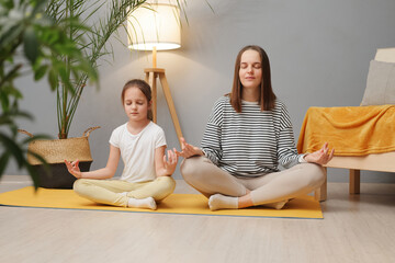 Family yoga. Beautiful young woman and her charming little daughter doing yoga together at home...