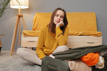 Bored Caucasian young adult woman packing suitcase at home taking too much attire waiting for her...