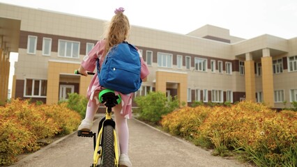 Little girl pupil with backpack riding on bike to elementary school lesson on schoolyard back view....