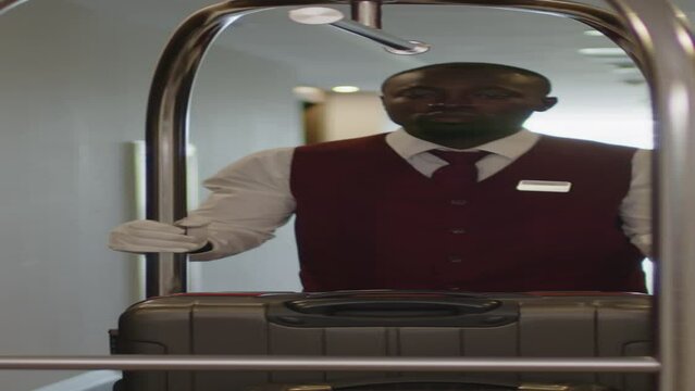 Vertical shot of young African American bellboy in uniform going through hotel hallway and carrying baggage on cart