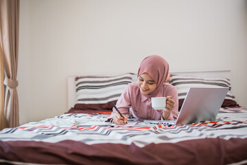 young woman in hijab works by writing while lying down and holding a cup in bed