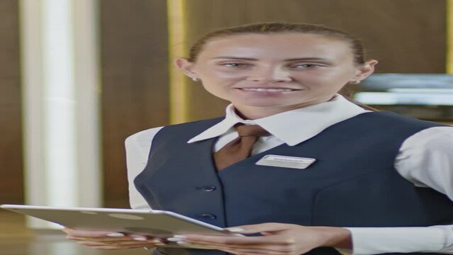 Vertical portrait shot of young female receptionist in uniform using digital tablet and then looking at camera with smile while standing in hotel lobby