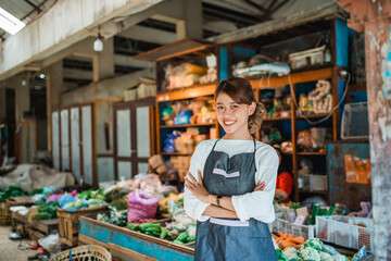 asian female vegetable seller standing in front of vegetables stall smiling with crossed arm - obrazy, fototapety, plakaty