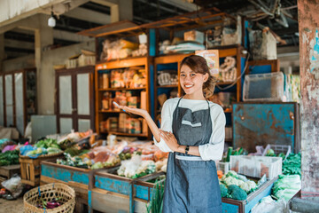 asian female vegetable seller standing in front of vegetables stall smiling with open hand