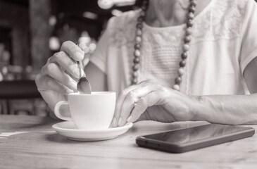 Black and white closeup on the hands of an elderly woman as she relaxes for a break at the cafe enjoying an espresso coffee, wooden table and mobile phone