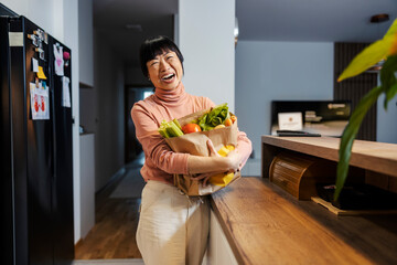 A cheerful middle-aged japanese woman holding groceries in her hands while standing in kitchen.