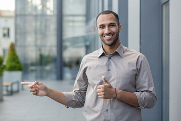 Confident young adult male smiling, giving thumbs up and pointing outdoors. Concept of approval,...