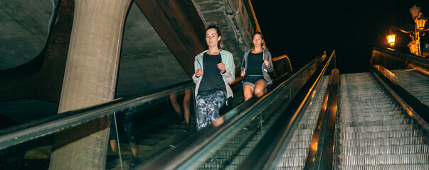 Two young women friends training running down escalator in the city at night