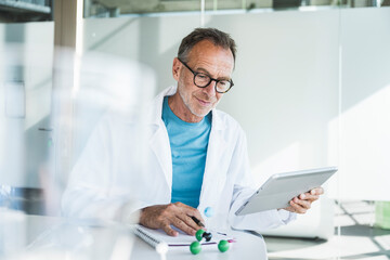 Smiling senior doctor using tablet PC and sitting with note pad at desk