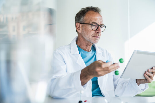 Man in lab coat holding molecular structure and using tablet PC at desk