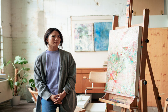 Confident artist standing by painting on easel in workshop