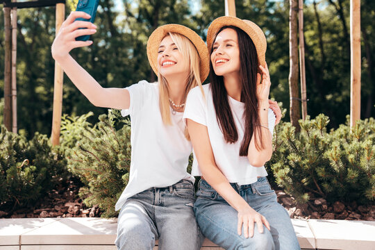 Two young beautiful smiling hipster female in trendy summer white t-shirt and jeans clothes. Carefree women posing in the street. Positive models having fun outdoors. Cheerful and happy. Take selfie