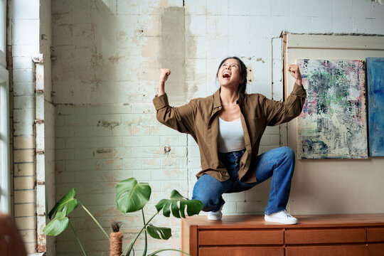 Excited painter shouting in joy squatting on cabinet by paintings at art studio